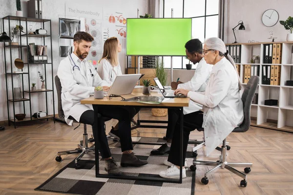 Team of multicultural medical specialists with thoughtful facial expression looking on monitor with green screen. Male and female doctors in lab coats having regular sessions in medical sphere.