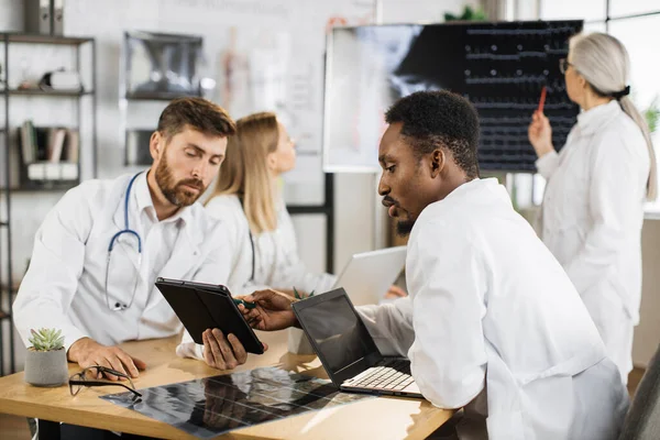 İki erkek doktor masada oturup tabletle dizüstü bilgisayar kullanırken kadın uzmanlar dijital monitörde röntgen taramasını inceliyor. Bir grup uluslararası tıp çalışanı toplantı odasında toplanıyor. .