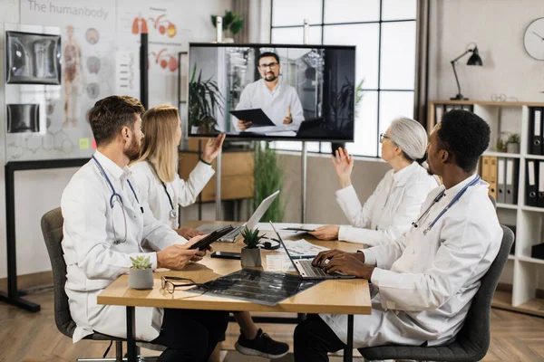 Çeşitli yaşlarda dört çok ırklı doktor toplantı odasında masada oturuyor ve dijital ekranda video görüşmesi sırasında erkek meslektaşlarını dinliyor. Uluslararası işbirliği ve tıp konsepti.