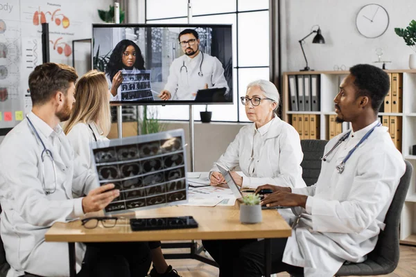 Teamwork of medical scientists examining x ray of patient lungs in modern clinic. Multicultural group of physicians in lab coats discussing MRI result with two colleagues during online video call.