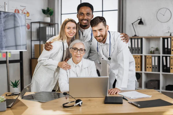 Group of multicultural medical workers in white lab coats hugging and looking at camera with smile in bright clinic boardroom. Effective teamwork and healthcare cooperation concept.