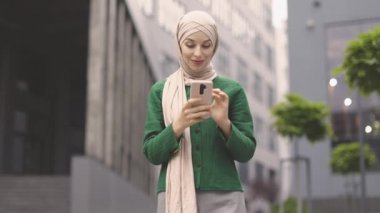 Modern ofis binasının yanında elinde akıllı telefonuyla duran güzel Müslüman kadının alçak açılı görüntüsü. Resmi giyimli ve tesettürlü genç iş kadını dışarıda iş e-postalarını kontrol ediyor..
