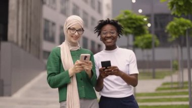 İki zarif kadın, bir beyaz Müslüman elbiseli ve Afrikalı Amerikalı, kameraya bakıp başparmaklarını kaldırarak gülümseyen arkadaşlar, şehir arka planında selfie portresi yapıyorlar..