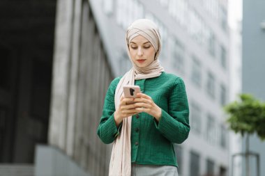 Modern ofis binasının yanında elinde akıllı telefonuyla duran güzel Müslüman kadının alçak açılı görüntüsü. Resmi giyimli ve tesettürlü genç iş kadını dışarıda iş e-postalarını kontrol ediyor..