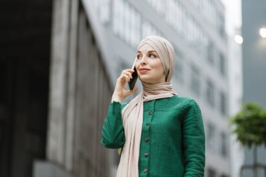 Yeşil ceketli ve tesettürlü Müslüman bir iş kadını sokakta dikilip telefonla konuşuyor. Kendine güvenen kadın girişimci telefonla konuşuyor, modern binaların arka planında.