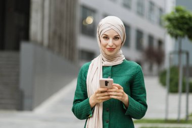 Arap iş kadını tesettürlü. Ofis binasının yanında dururken akıllı telefon kullanıyor. Teknoloji, iş ve modern yaşam tarzları kavramı. Başarılı bir kadın kariyeri..