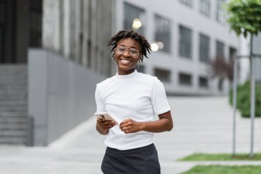 Modern iş merkezinin yanında elinde akıllı telefonuyla gündelik kıyafetlerle dikilen Afro-Amerikan kadın portresi. Başarılı iş kadını modern şehrin arka planında poz veriyor..