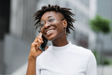 Modern şehirde yürürken cep telefonuyla konuşan Afro-Amerikan iş kadınının portresi. Şık güneş gözlüklü güzel kız iş merkezinde yürürken akıllı telefonla konuşuyor.