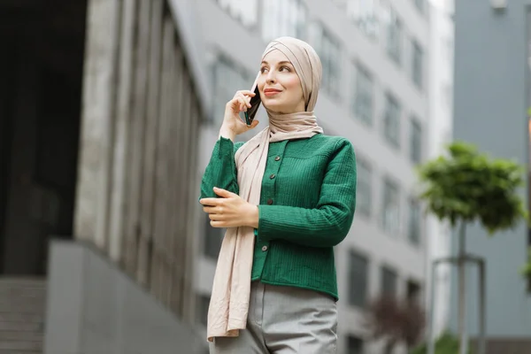 Muslim business woman in green jacket and hijab standing on street and talking on mobile. Confident female entrepreneur having working conversation on phone, outdoor on background of modern buildings