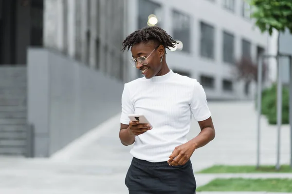 Front view of pleasant business african woman using modern smartphone while standing outdoors. Young African American lady typing messages on mobile outside office.