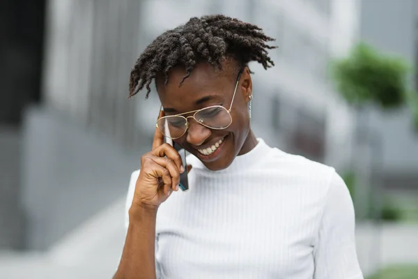 Portrait of african american business woman talking on cell phone as she walking through the modern city. Pretty girl with stylish sunglasses talking on smart phone while walking on business center