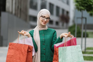 Alışveriş konsepti. Çekici, neşeli Müslüman kadının yakın portresi, renkli alışveriş torbalarıyla kameraya gülümsüyor, bulanık şehir arka planında poz veriyor..