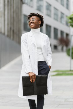 Hastanenin dışında gülümseyen çekici bir Afrikalı kadın doktor. Modern üniversite binası veya kliniği önünde poz veren beyaz önlüklü bayan tıp öğrencisinin tam boy portresi.