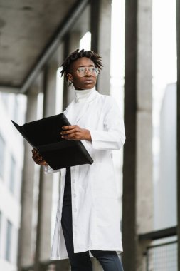 Beyaz önlüklü hoş, Afro-Amerikalı kadın doktoru yakından izleyin. Elinde bir pano, modern kliniğin merdivenlerinde duruyor, gülümsüyor ve kameraya bakıyor..