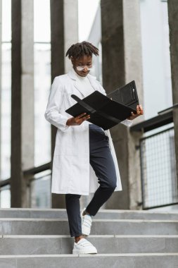 Dosyalama panosuyla modern binanın merdivenlerinde duran, yetenekli, genç Afrikalı kadın sağlık görevlisi..