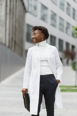 Yüzünde dostane bir klasörle modern klinik ya da kampüsün dışında duran, gülümseyen, Afro-Amerikan doktor ya da beyaz önlüklü öğrencinin açık havada çekilmiş fotoğrafı..