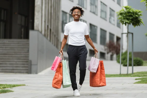 Horizontal outdoor shot of cheerful African girl in stylish fashionable clothes, doing shopping in city. African American female walking with shopping bags on the background of modern office buildings