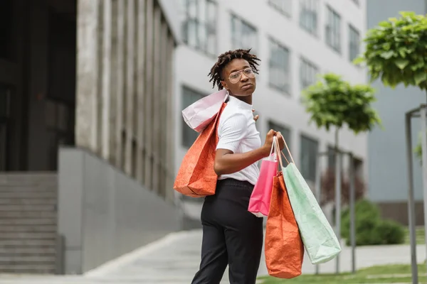 Shopping, city lifestyle concept. Outdoor bright portrait of young attractive African American woman, posing on the background of office buildings of modern city, showing colorful shopping bags.