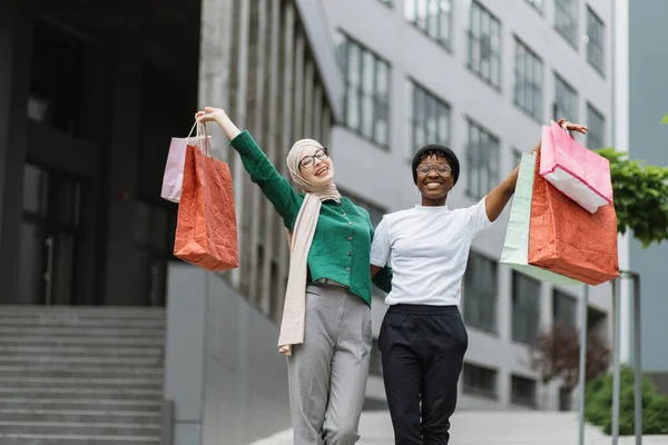 City shopping concept. Outdoor urban lifestyle portrait of two diverse young women, African and Muslim, holding colorful shopping bags while walking in the modern city.