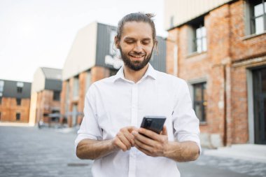 Beyaz tişörtlü, sakallı, yakışıklı, gülümseyen bir iş adamı şehirde dolaşıyor ve akıllı telefon kullanıyor. Mutlu adam dışarıda dikilip dijital cihazlarla e-postaları kontrol ediyor. Yaşam tarzı kavramı.