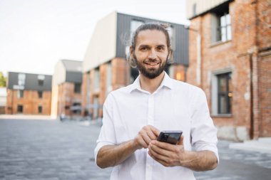Beyaz tişörtlü, sakallı, yakışıklı, gülümseyen bir iş adamı şehirde dolaşıyor ve akıllı telefon kullanıyor. Mutlu adam dışarıda dikilip dijital cihazlarla e-postaları kontrol ediyor. Yaşam tarzı kavramı.