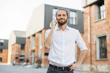 Bir telefon görüşmesi yapan gülümseyen genç adamın portresi. Yakışıklı, sakallı, siyah saçlı adam açık havada akıllı telefon kullanıyor..