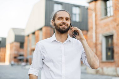 Bir telefon görüşmesi yapan gülümseyen genç adamın portresi. Yakışıklı, sakallı, siyah saçlı adam açık havada akıllı telefon kullanıyor..