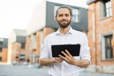 Beyaz gömlekli, sakallı yakışıklı iş adamı şehirde dolaşıyor ve tablet kullanıyor. Mutlu adam dışarıda dikiliyor ve dijital cihazlarla e-postaları kontrol ediyor. Yaşam tarzı kavramı.