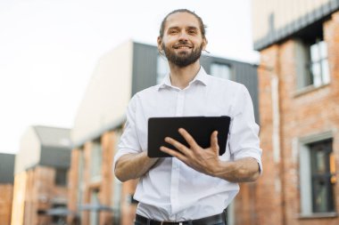 Beyaz gömlekli, sakallı yakışıklı iş adamı şehirde dolaşıyor ve tablet kullanıyor. Mutlu adam dışarıda dikiliyor ve dijital cihazlarla e-postaları kontrol ediyor. Yaşam tarzı kavramı.