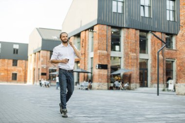 Beyaz gömlekli bir iş adamı akıllı telefondan konuşuyor. Profesyonel işadamı cep telefonu kullanıyor, gülümsüyor ve şehirdeki ofis binasının yakınında kahve içiyor..