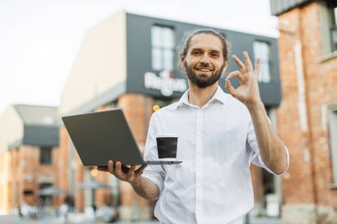 Yakışıklı beyaz adamın portresi kağıt bardakta taze içecek içerken partneriyle sohbet ederken internette dizüstü bilgisayarla sokakta yürürken kameraya bakıp işaret veriyor..