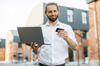 İnternetten alışveriş yapmak için laptop ve kredi kartı kullanan beyaz gömlekli yakışıklı bir adam. Sakallı işadamı sokakta dikiliyor ve alışveriş yapıyor..