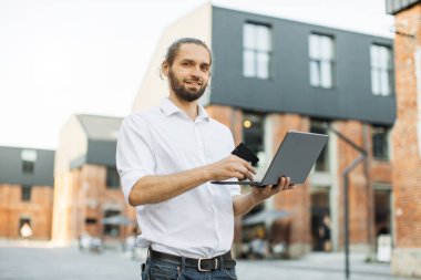 Ofis binasının önünde dikilen şık sakallı adam internetten alışveriş yapıyor. Resmi olarak beyaz adam alışveriş için kredi kartı ve laptop kullanıyor..