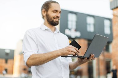 Ofis binasının önünde dikilen şık sakallı adam internetten alışveriş yapıyor. Resmi olarak beyaz adam alışveriş için kredi kartı ve laptop kullanıyor..