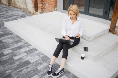 İnternetten alışveriş yapmak için laptop ve kredi kartı kullanan beyaz gömlekli ve siyah pantolonlu çekici bir kadın. Mola sırasında merdivenlerde oturan ve alışveriş yapan pozitif iş kadını..