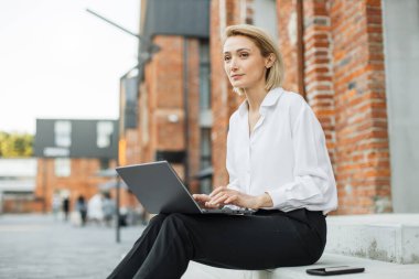 Modern dizüstü bilgisayarla ofis binasının yanındaki merdivenlerde oturan şık bir iş kadını. Gülümseyen kadın serbest yazar tatilde en sevdiği işi yapıyor..