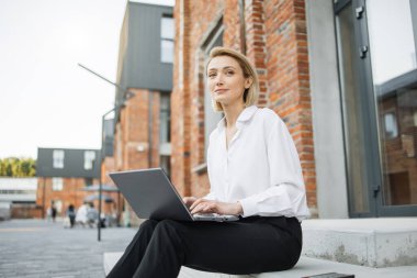 Modern dizüstü bilgisayarla ofis binasının yanındaki merdivenlerde oturan şık bir iş kadını. Gülümseyen kadın serbest yazar tatilde en sevdiği işi yapıyor..