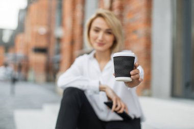Kahveye odaklan. Gülümseyen iş kadını elinde bir fincan içecek ve cep telefonuyla konuşuyor. Arka planda modern şehir hayatı. Beyaz gömlekli kadın merdivenlerde oturuyor ve cep telefonuyla konuşuyor..