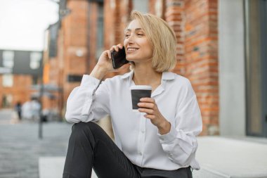 Moda kıyafetleri içinde şık giyinen, akıllı telefondan konuşan ve şehir caddesindeki merdivenlerde otururken hazır kahve içen neşeli bayanı kapatın..