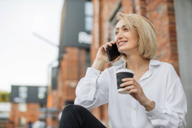 Moda kıyafetleri içinde şık giyinen, akıllı telefondan konuşan ve şehir caddesindeki merdivenlerde otururken hazır kahve içen neşeli bayanı kapatın..
