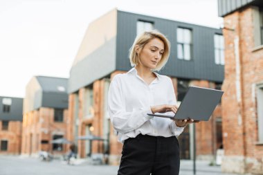 Modern dizüstü bilgisayarını ofis binasının yakınındaki sokakta yürürken kullanan şık bir iş kadını. Gülümseyen kadın serbest yazar tatilde en sevdiği işi yapıyor..