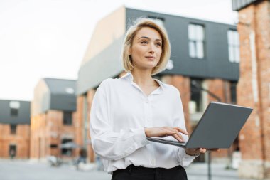 Düşük açı. Beyaz gömlekli ve pantolonlu başarılı bir iş kadını şehir sokaklarında dikiliyor ve modern dizüstü bilgisayarda daktilo kullanıyor. Uzak çalışma için taşınabilir bilgisayar kullanan çekici bir serbest yazar