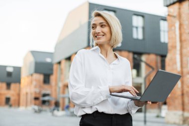 Düşük açı. Beyaz gömlekli ve pantolonlu başarılı bir iş kadını şehir sokaklarında dikiliyor ve modern dizüstü bilgisayarda daktilo kullanıyor. Uzak çalışma için taşınabilir bilgisayar kullanan çekici bir serbest yazar