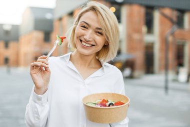 Hoş bir gülümsemesi olan genç bir kadın öğle arasında şehirde yürürken taze sebze salatası yiyor..