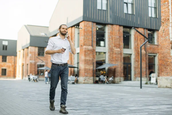 Beyaz gömlekli bir iş adamı akıllı telefondan konuşuyor. Profesyonel işadamı cep telefonu kullanıyor, gülümsüyor ve şehirdeki ofis binasının yakınında kahve içiyor..