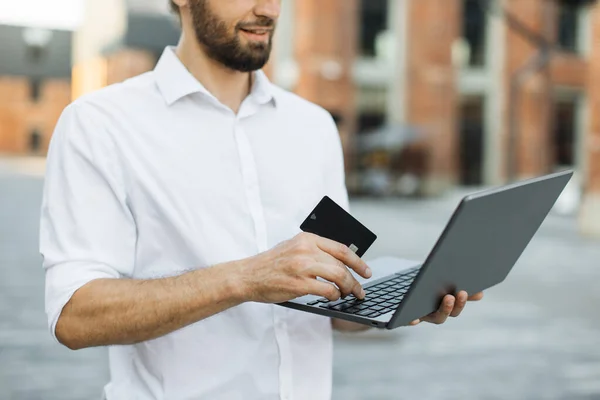 Cropped view. Online payment concept. Confident young businessman in formal wear, stands outside at office building, uses laptop and credit bank card doing pay for air flight.