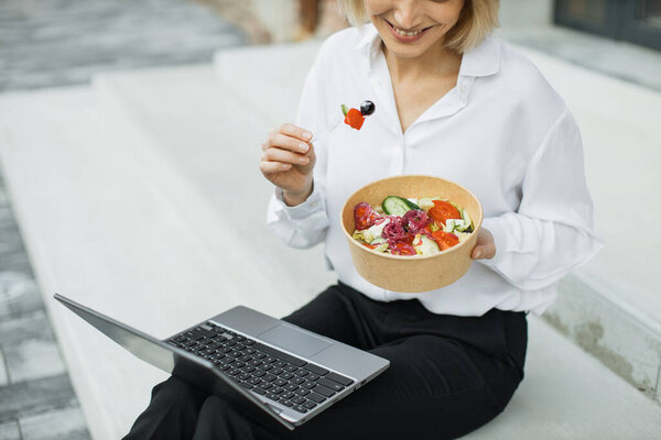 Close up view of hand of businesswomann having a vegetables salad for lunch, healthy eating sitting on stairs using modern laptop, lifestyle concept, unrecognizable person.