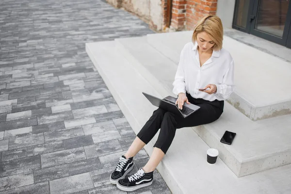 İnternetten alışveriş yapmak için laptop ve kredi kartı kullanan beyaz gömlekli ve siyah pantolonlu çekici bir kadın. Mola sırasında merdivenlerde oturan ve alışveriş yapan pozitif iş kadını..