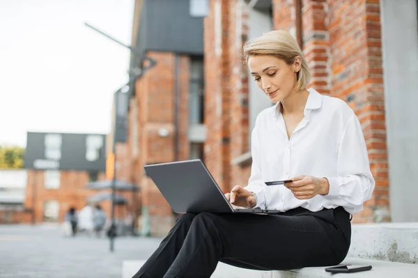 Online payment concept. Confident young businesswoman in formal wear, sitting on stairs outside office building, uses laptop and credit bank card doing pay for air flight.