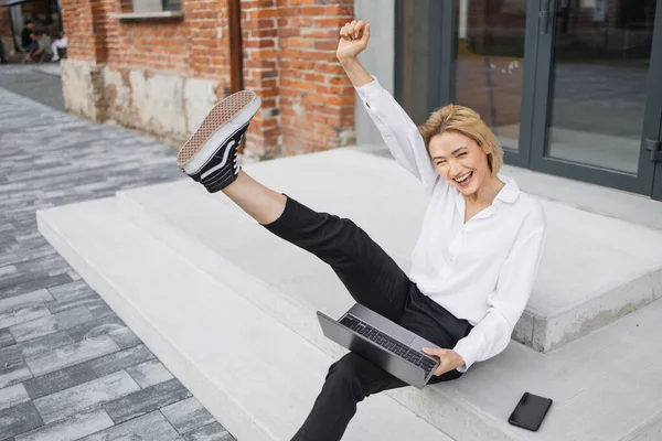 Happy satisfied caucasian business woman, relaxing while sitting on the steps of a office building rejoicing with laptop doing online order after shopping. Success and happy news concept.